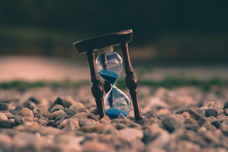 Daily Walk Devotion hourglass on pebbles by a lake, mindfulness reminder.