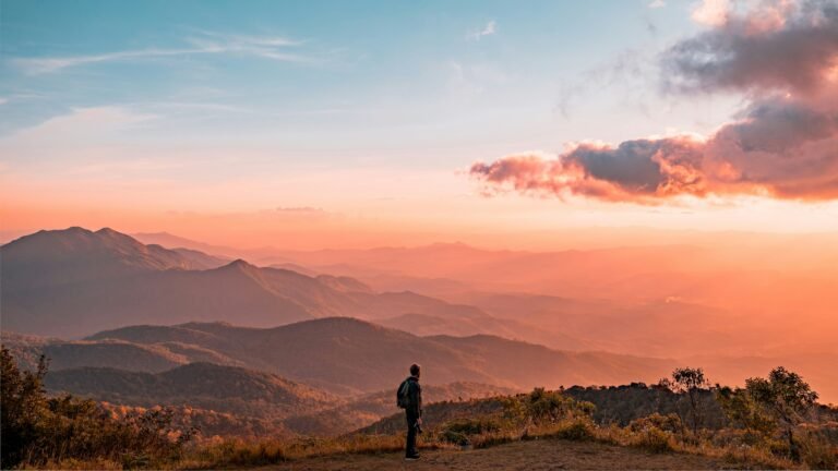Person hiking on a mountainside at sunset with orange-pink sky and distant misty ranges.