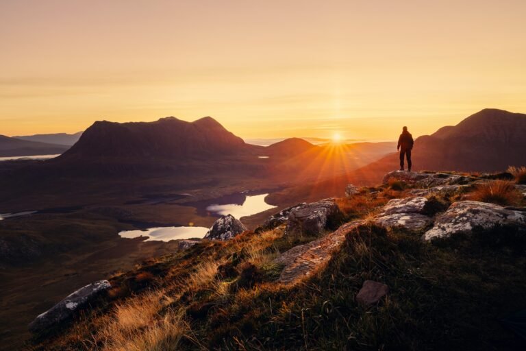 Hiker on a rocky mountaintop at sunset over a valley and lake.