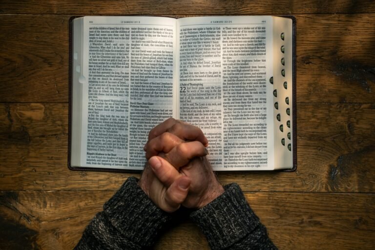 Praying hands over open Bible on a wooden table.