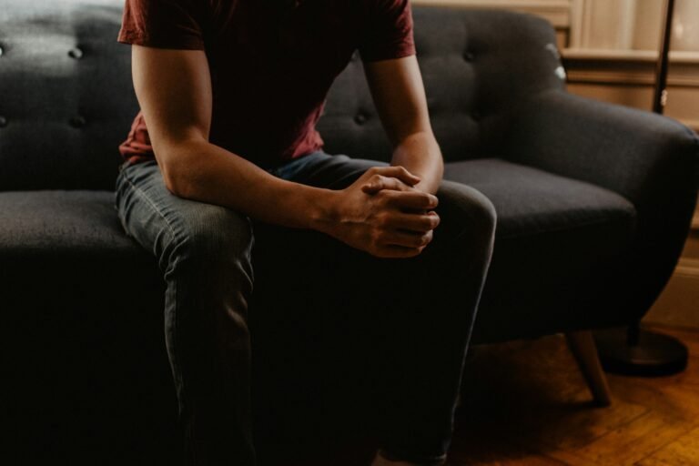Person sitting on a couch with hands clasped in lap, indoors.
