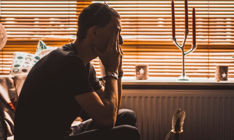 Man sits with his head in hands in a dimly lit room with blinds and a microphone nearby.