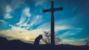 Silhouette of a person kneeling beside a cross at sunset.