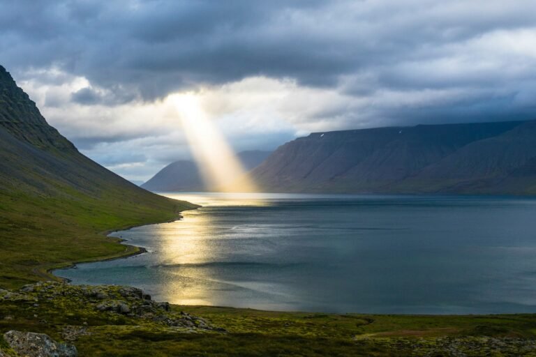 Sunbeam breaking through clouds over a tranquil fjord landscape with mountains and water.