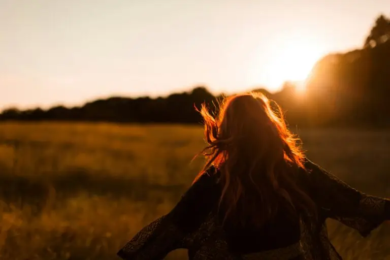 Person raising arms in a sunny field at sunset.