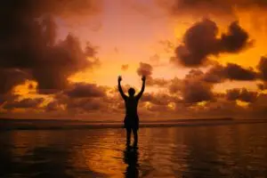 Person standing in shallow water with arms raised during a sunset.