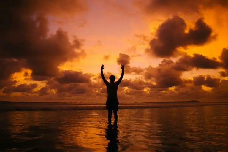Person standing in shallow water with arms raised during a sunset.
