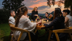 People gathered around a table outdoors at sunset sharing a meal and drinks.