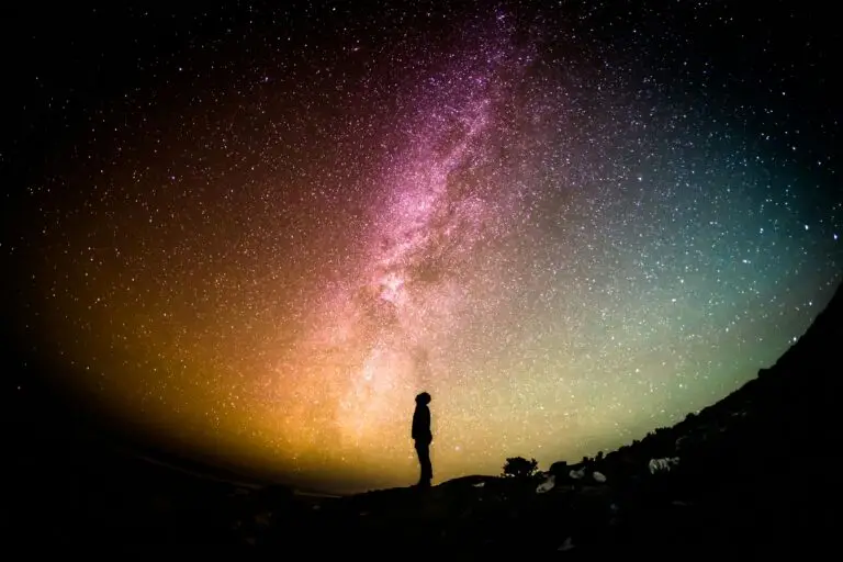 Silhouette of a person under a colorful Milky Way night sky over a hill.