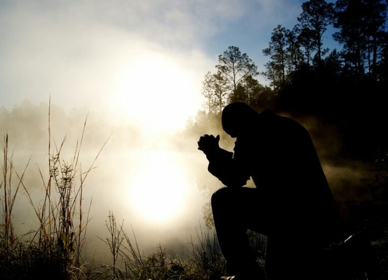 Silhouette of a person kneeling by a misty lake at sunrise, hands clasped in prayer.