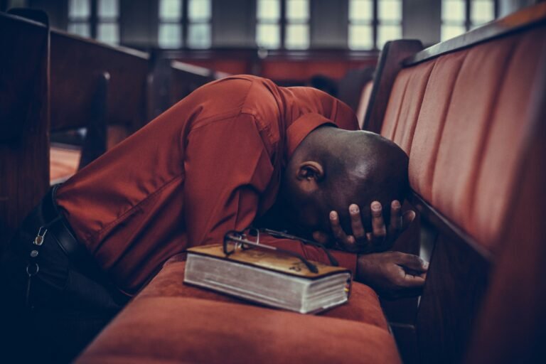 Person praying with head down on church pews beside a Bible.